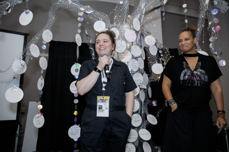 Meredith Hutchison speaking into a microphone beside a collaborator in front of an interactive installation of handwritten notes on a silver tree.