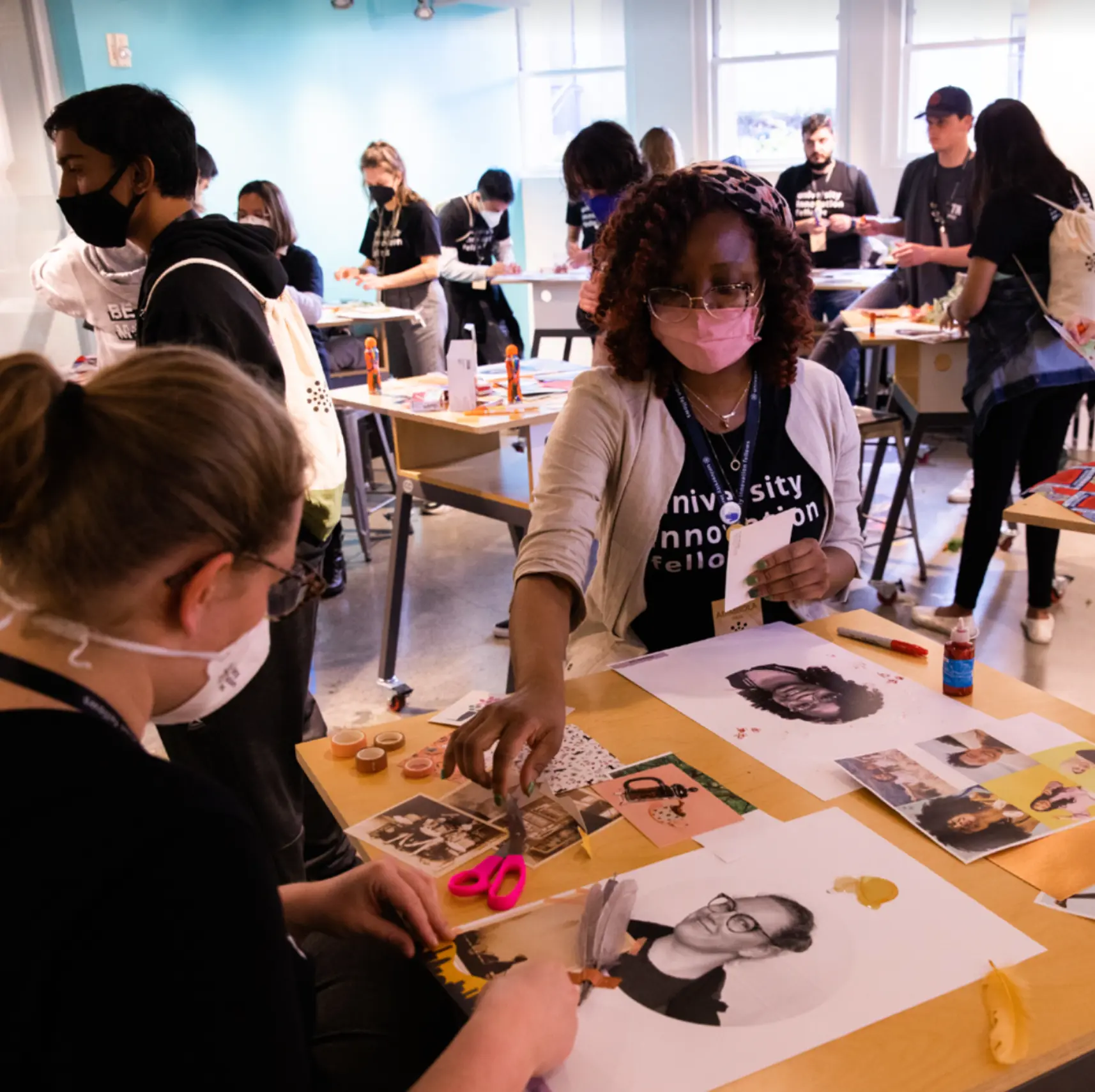 Masked participants gathered around a workshop table cutting and arranging printed portrait collages with scissors and glue.