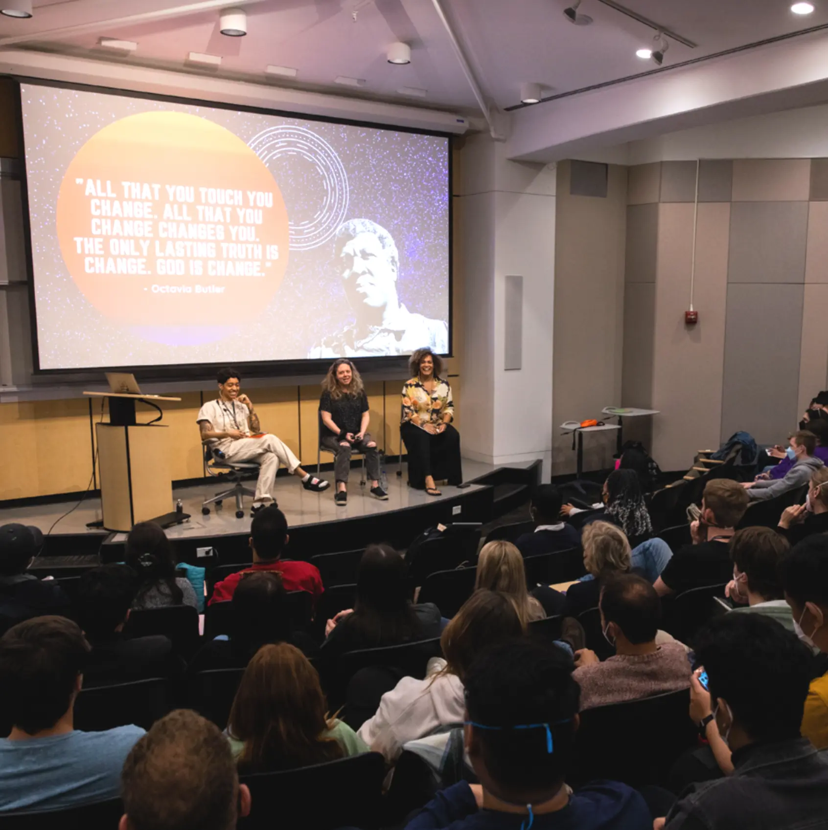 Panel discussion in a lecture hall with an Octavia Butler quote about change projected large on the screen behind three seated speakers.